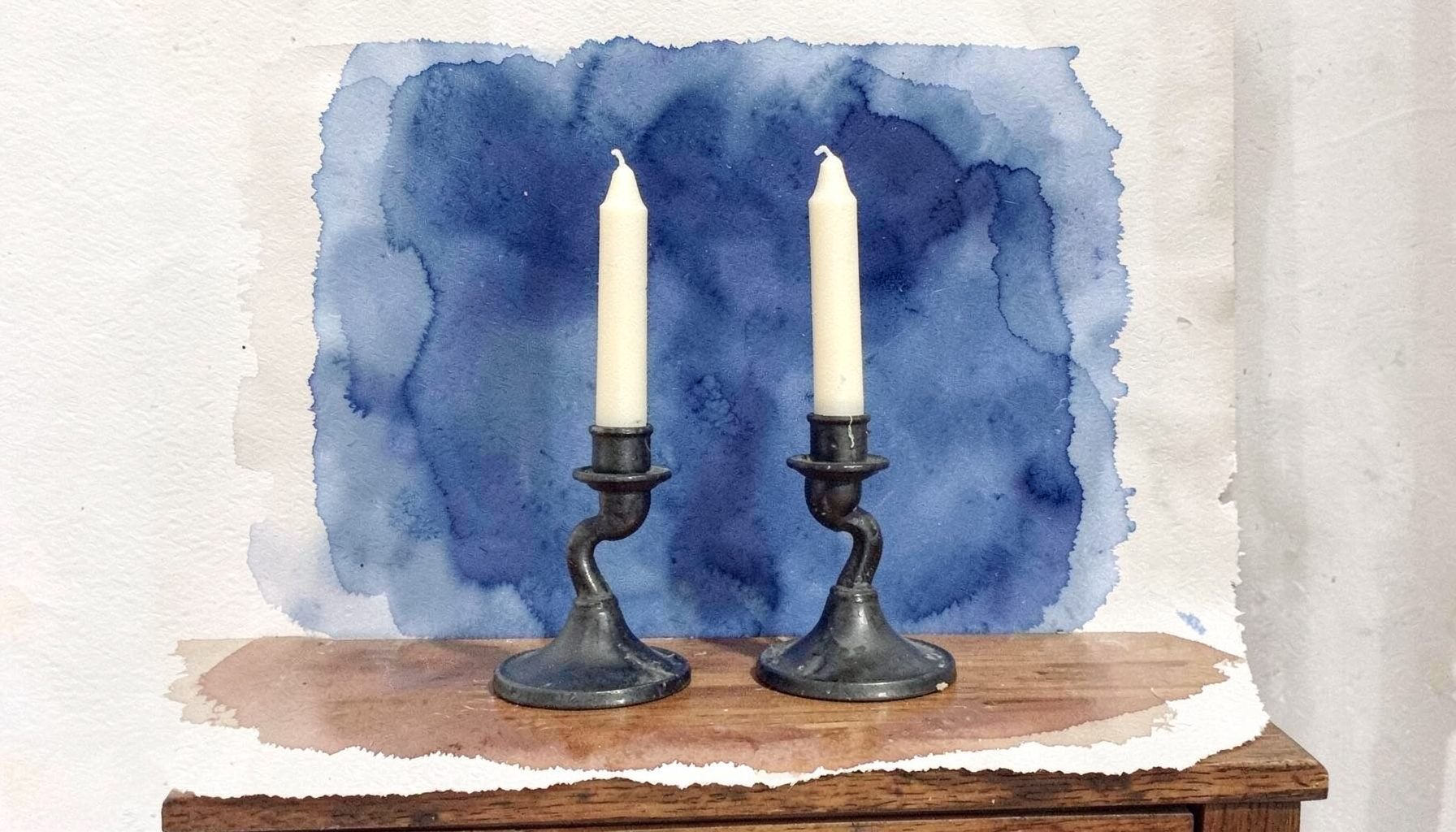 A pair of wrought iron candleholders with a dark matte finish, holding cream taper candles on a wooden sideboard